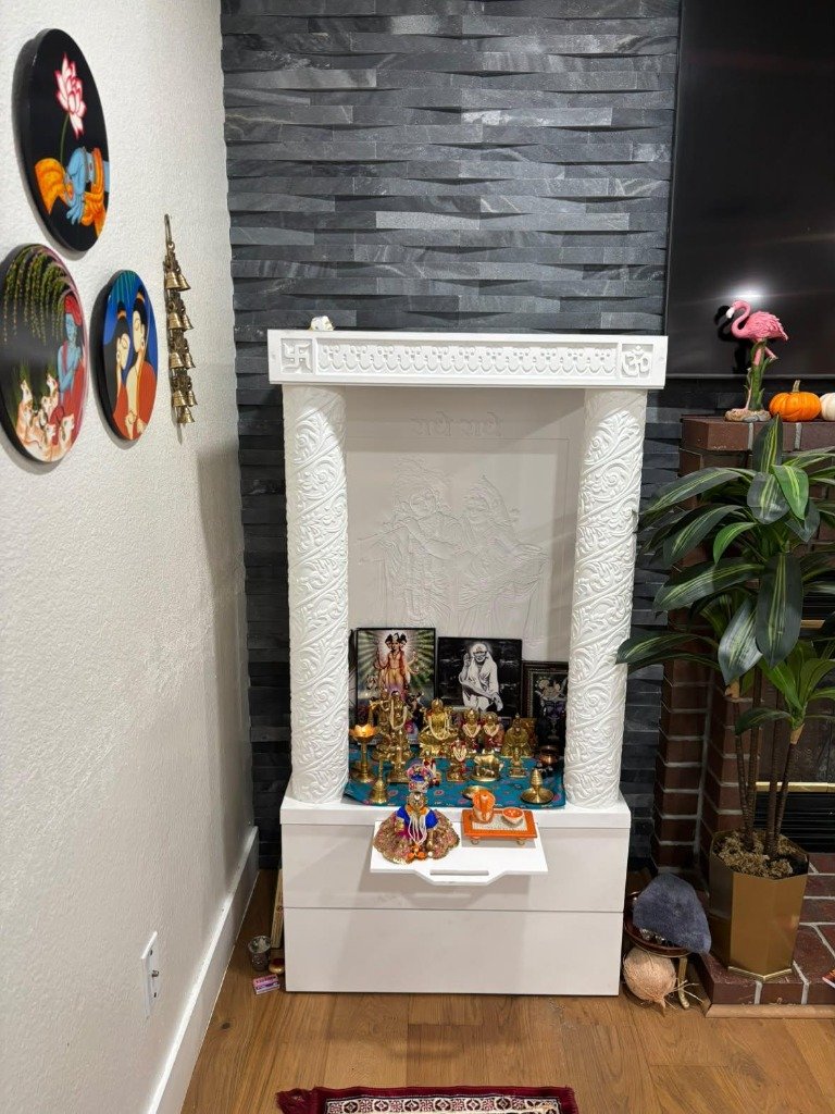 Intricately carved white wooden pooja mandir with Radha Krishna relief, brass deities, and brass bells installed in an NRI home abroad